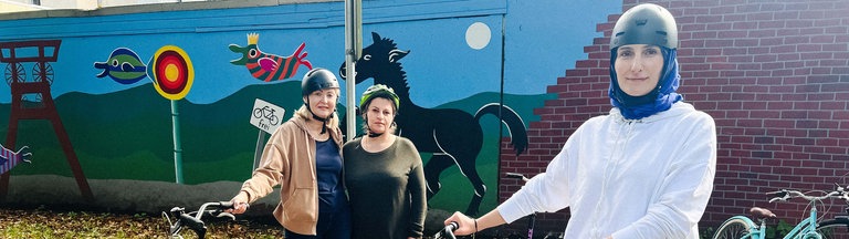 Drei Frauen mit Helm stehen neben zwei Fahrrädern auf der Straße.