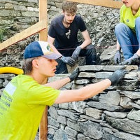 Der Ehrenamtliche Christoph Hustedt stapelt konzentriert längliche Steine aufeinander. Es soll eine stabile Steinmauer entstehen. Er hat Handschuhe und Arbeitskleidung an. Zwei andere Jugendliche schauen ihm dabei zu.