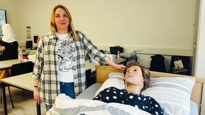Eine Frau steht neben einem Krankenbett, in dem eine lebensgroße Puppe liegt