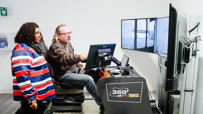 Ein Mann sitzt an einem Fahrsimulator, neben ihm steht eine Frau
