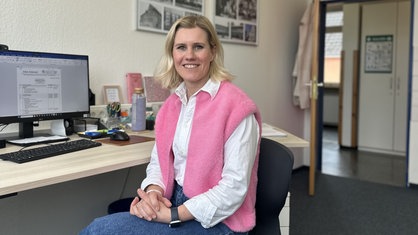 Eine Frau mit mittellangen blonden Haaren und einer rosafarbenen Weste lächelt. Im Hintergrund ein Büro