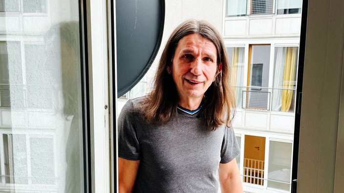 Willi Lehmann steht auf dem Balkon seiner Kölner Wohnung, lehnt am Türrahmen und lächelt.