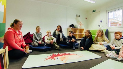 Eine Frau sitzt mit einem Stofftierigel auf dem Boden. Neben ihr sitzen mehrere Kinder, die ihr zuhören.