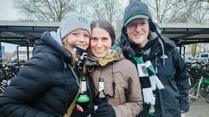 Zwei Frauen und ein Mann stehen in Winterjacken eng nebeneinander und lachen in die Kamera. Sie tragen jeweils einen schwarz-weiß-grünen, gehäkelten Becherhalter, in dem Bierflaschen stecken.