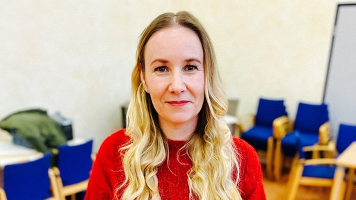 Ein Frau mit langen blonden Haaren und einem roten Pullover schaut direkt in die Kamera.