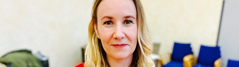 Ein Frau mit langen blonden Haaren und einem roten Pullover schaut direkt in die Kamera.