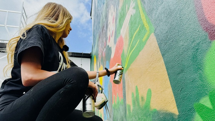 Eine junge Frau mit langen Haaren sprayt an einer Gebäudewand.