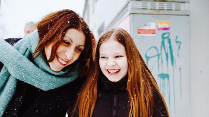 Ida und Nanis stehen draußen in Winterjacken vor einem Stromkasten. Ihre Haare sind von Schneeflocken bedeckt und beide lachen. 