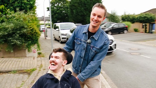 Hospizdienstmitarbeiter Jan Meyer schiebt Hubertus Kaiser in einem Rollstuhl über einen Bürgersteig.