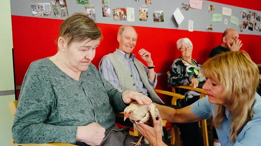 Birgit Brammertz hält ein Huhn. Neben ihr sitzt eine Seniorin, die das Huhn streichelt.