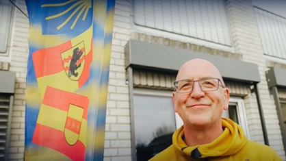 Thomas Fischer, Glatze, Brille und gelber Pulli, steht hinter seinem Fahhrad auf einer Terrasse, am Fahrrad ist eine Flagge mit angebracht auf den Flaggen von mehreren Nationen zu sehen sind
