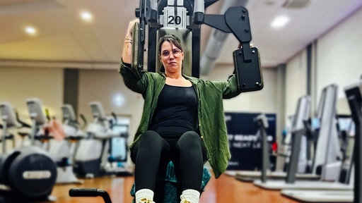 Angelina Schirmer sitzt auf einem Butterfly-Gerät im Fitnessstudio. Ihren rechten Arm am Gerät drückt sie nach vorne, der linke bleibt hinten.