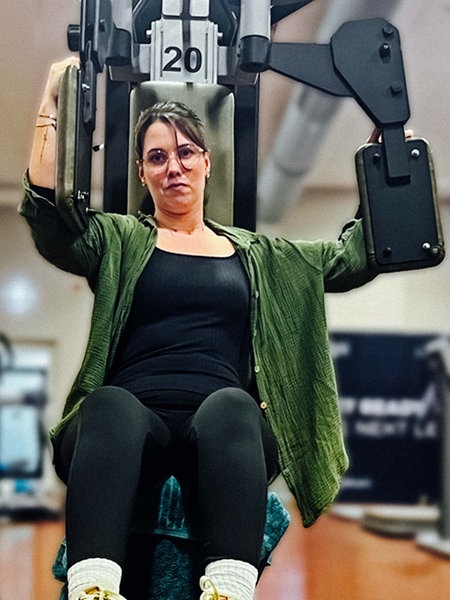 Angelina Schirmer sitzt auf einem Butterfly-Gerät im Fitnessstudio. Ihren rechten Arm am Gerät drückt sie nach vorne, der linke bleibt hinten.