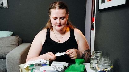 Eine Frau sitzt an einem Tisch. Auf dem Tisch liegen mehrere Verpackungen von Medikamenten. In ihren Händen hält sie einen Tablettenblister.