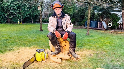 Luis Hantschel, ein junger Mann im Arbeitsanzug, orangem Schutzhelm und Handschuhen, sitzt auf einem gesägten Holzstumpf. Vor ihm links liegt eine gelbe Kettensäge.