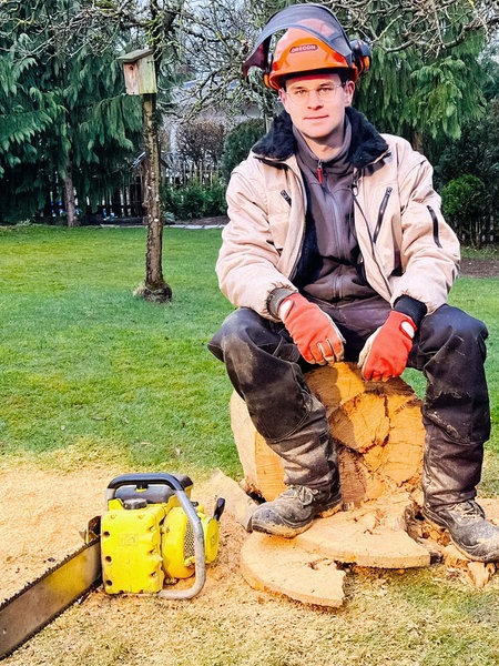 Luis Hantschel, ein junger Mann im Arbeitsanzug, orangem Schutzhelm und Handschuhen, sitzt auf einem gesägten Holzstumpf. Vor ihm links liegt eine gelbe Kettensäge.