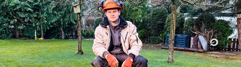 Luis Hantschel, ein junger Mann im Arbeitsanzug, orangem Schutzhelm und Handschuhen, sitzt auf einem gesägten Holzstumpf. Vor ihm links liegt eine gelbe Kettensäge.