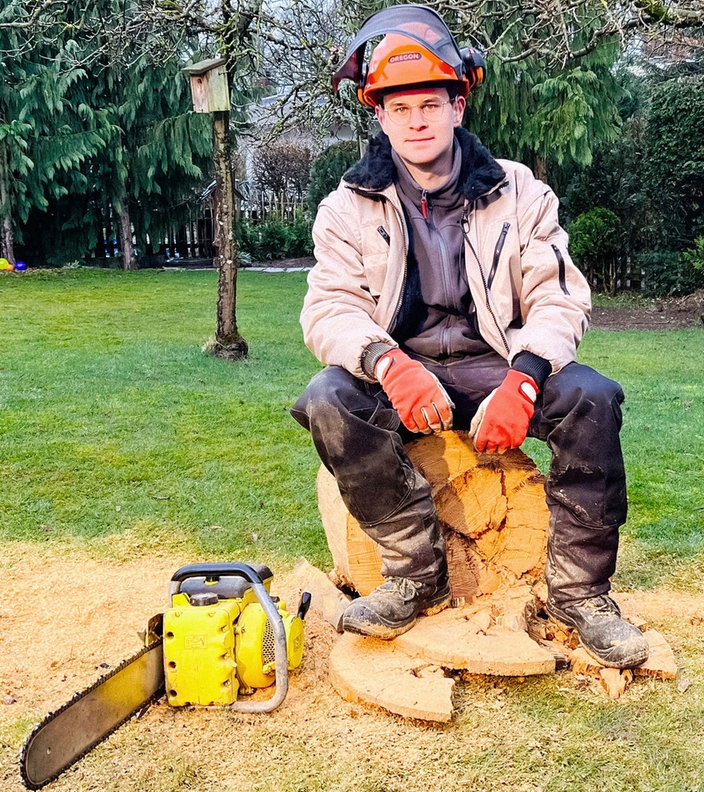 Luis Hantschel, ein junger Mann im Arbeitsanzug, orangem Schutzhelm und Handschuhen, sitzt auf einem gesägten Holzstumpf. Vor ihm links liegt eine gelbe Kettensäge.