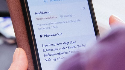 Nahaufnahme eines in einer Hand gehaltenen Smartphones, das eine Pflegedokumentation auf dem Display zeigt.
