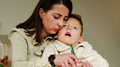 Eine Mutter hat ihren Sohn, der einen Gendefekt hat, auf dem Schoß und malt zusammen mit ihm am Tisch.
