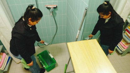 Gabriela steht in einem grün gefliesten Bad und füllt Wasser in einen Eimer.