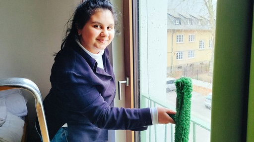 Gabriela, eine junge brünette Frau im schwarzen Pullover, steht vor einem Fenster und hält einen nassen grünen Wischer gegen die Scheibe.