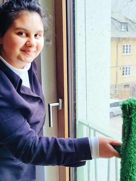 Gabriela, eine junge brünette Frau im schwarzen Pullover, steht vor einem Fenster und hält einen nassen grünen Wischer gegen die Scheibe.
