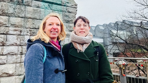 Elke, eine Frau mit langen blonden Haaren, und Jenny, kurze braune Haare und Brille, stehen vor einer Mauer und lächeln in die Kamera.
