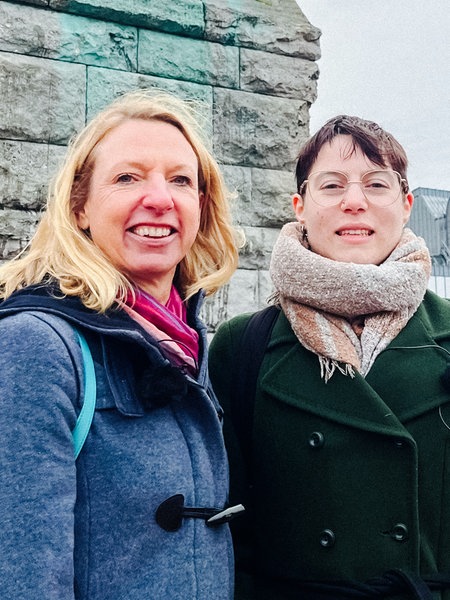 Elke, eine Frau mit langen blonden Haaren, und Jenny, kurze braune Haare und Brille, stehen vor einer Mauer und lächeln in die Kamera.