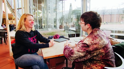 Elke und Jenny sitzen sich an einem Tisch gegenüber und unterhalten sich.