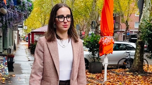 Simin Heuser, eine junge brünette Frau in einem hellen, karierten Jackett und einem weißen T-Shirt, läuft auf einem Gehweg unter begrünten Bäumen.
