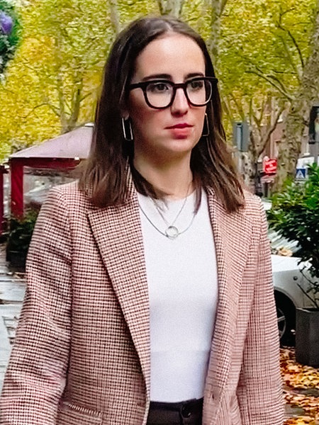 Simin Heuser, eine junge brünette Frau in einem hellen, karierten Jackett und einem weißen T-Shirt, läuft auf einem Gehweg unter begrünten Bäumen.