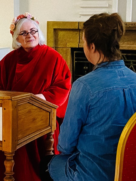 Marie-Luise O'Byrne-Brandl sitzt hinter einem braunen Holzpult. Sie trägt ein rotes Gewand und einen Blumenkranz auf dem Kopf.