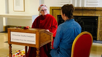 Marie-Luise O'Byrne-Brandl sitzt hinter einem braunen Holzpult. Sie trägt ein rotes Gewand und einen Blumenkranz auf dem Kopf.