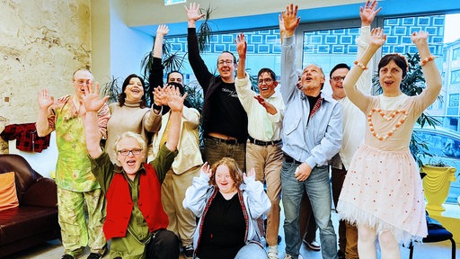 Eine Gruppe Menschen, zum Teil in Kostümen, winkt in die Kamera