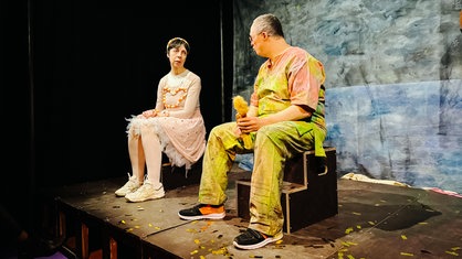 Eine Frau und ein Mann sitzen in bunten Kostümen auf einer Theaterbühne und agieren miteinander