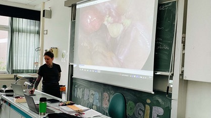 Ulrike Kuhlmann steht am Pult im Klassenraum. Hinter ihr auf einer Leinwand läuft ihr OP-Video.