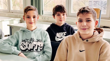 Drei Schüler sitzen nebeneinander im Klassenraum an einem Tisch. Sie lächeln.