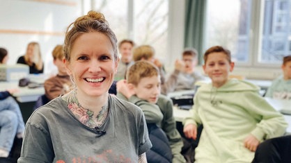 Rechts sitzt Ulrike Kuhlmann. Sie lächelt. Hinter ihr füllen Schüler den Klassenraum.