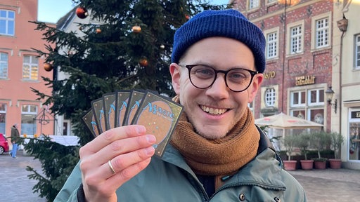 Felix Schneegans, blaue Mütze, schwarz-umrandetet Brille, orangener Schal, lächelt, in der rechten Hand hält er sechs Karten hoch, auf denen in blauer Schrift "Magic" steht.
