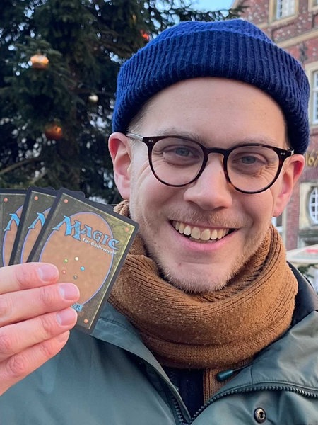 Felix Schneegans, blaue Mütze, schwarz-umrandetet Brille, orangener Schal, lächelt, in der rechten Hand hält er sechs Karten hoch, auf denen in blauer Schrift "Magic" steht.