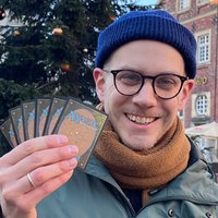 Felix Schneegans, blaue Mütze, schwarz-umrandetet Brille, orangener Schal, lächelt, in der rechten Hand hält er sechs Karten hoch, auf denen in blauer Schrift "Magic" steht.