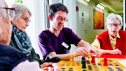 Sebastian Horstmann sitzt mit drei Seniorinnen an einem Tisch und spielt "Mensch ärgere dich nicht".