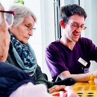 Sebastian Horstmann sitzt mit drei Seniorinnen an einem Tisch und spielt "Mensch ärgere dich nicht".