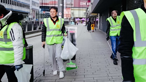 Eiichiro Kawasaki in einer Warnweste verteilt in der Düsseldorfer Innenstadt Arbeitshandschuhe an seine Helfer. Einer davon steht links von ihm.