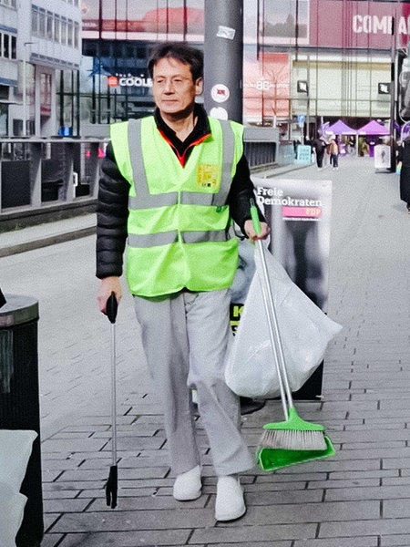 Eiichiro Kawasaki in einer Warnweste verteilt in der Düsseldorfer Innenstadt Arbeitshandschuhe an seine Helfer. Einer davon steht links von ihm.