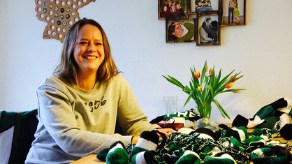 Eine Frau mit blonden Haaren sitzt an einem Tisch und lacht herzlich. Auf dem Tisch liegt schwarz-weiß-grüne Häkelwolle