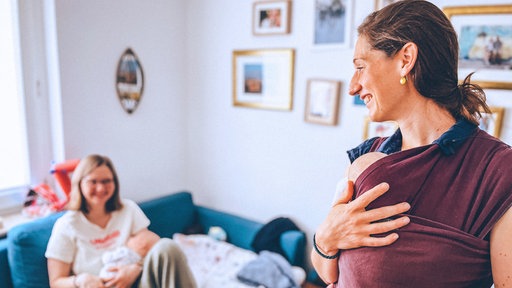 Links sitzt Mutter Greta auf der Couch, ein Baby im Arm. Rechts steht Anna van der Luijt, trägt ebenfalls ein Baby und lächelt.