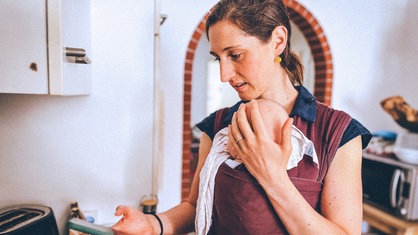 Anna van der Luijt steht in der Küche, ein Baby in einer Trage, das Handy in der Hand.