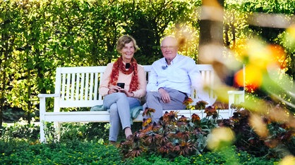 Karin Kretschmer und Friedbert Kretschmer sitzen im Grünen auf einer Bank und lächeln.